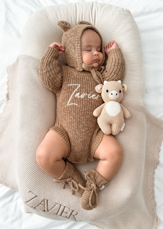 Baby in a brown knitted outfit with bear ears, lying on a white blanket with 'Zavier' embroidered.