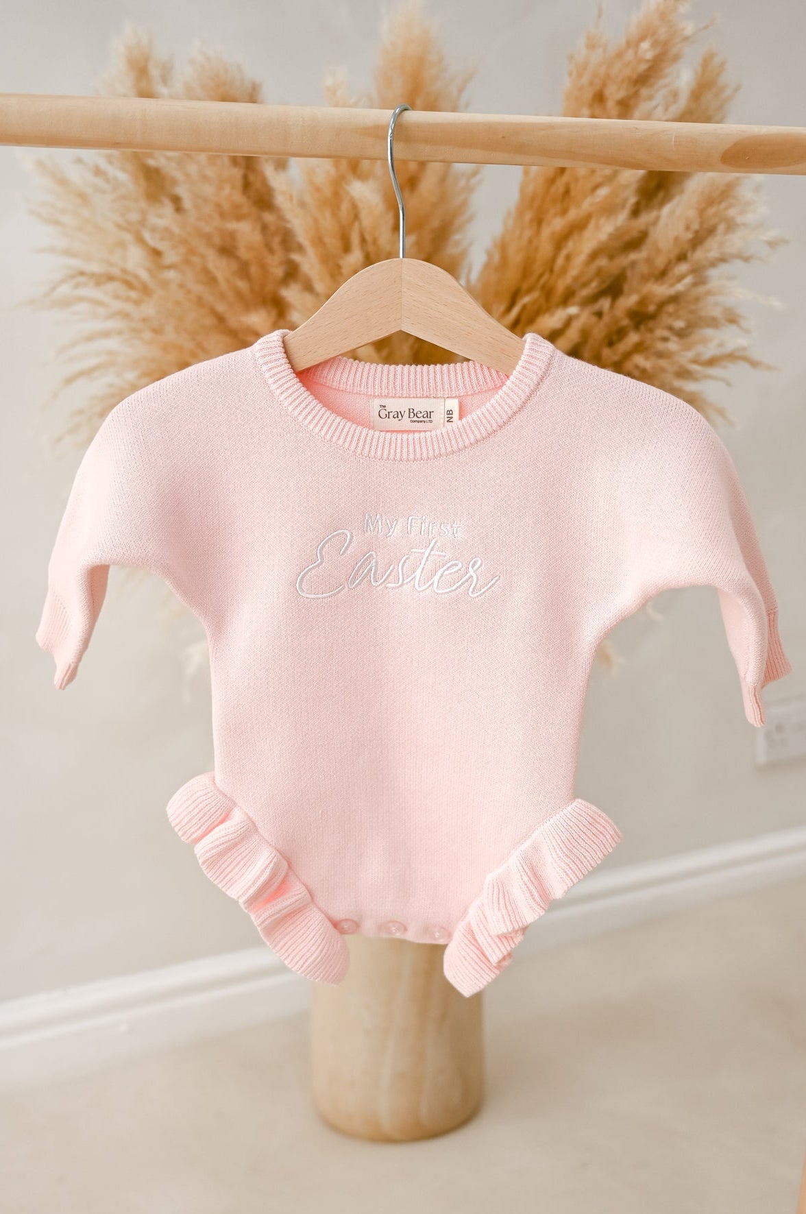 Pink baby onesie with ruffled legs and bottom hanging on a wooden hanger against a neutral background