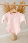 Pink baby onesie with ruffled legs and bottom hanging on a wooden hanger against a neutral background